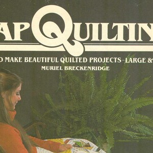 May include: Book cover with the title "Lap Quilting: How to Make Beautiful Quilted Projects - Large & Small" by Muriel Breckenridge. The cover features a woman sitting in a rocking chair with a quilt in her lap, and a fern plant in the background.