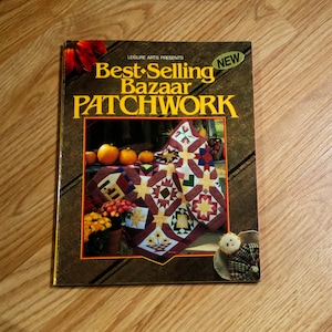 May include: A book titled "Best-Selling Bazaar Patchwork" with a "NEW" sticker. The cover features a patchwork quilt with star and floral designs, pumpkins, and fall foliage. The book is on a wooden surface.