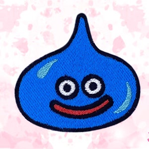 Può includere: Toppa di slime blu ricamata con contorno nero. La toppa presenta due occhi bianchi e una bocca rossa sorridente. Il design ricorda un popolare personaggio dei videogiochi. Lo sfondo è un disegno ad acquerello rosa chiaro.