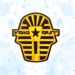May include: Embroidered patch featuring a stylized pharaoh's mask in yellow and black, with the text "VOID OUT" and a star. The patch has a textured appearance and is likely designed for clothing or accessories.