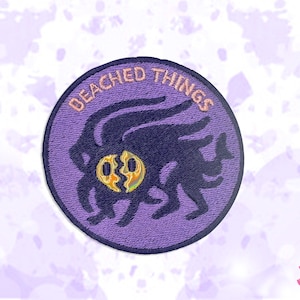 Könnte beinhalten: Ein kreisförmiger, lila gestickter Aufnäher mit den Worten "BEACHED THINGS" in einem Halbkreis oben. Der Aufnäher zeigt eine dunkelblaue, abstrakte Kreatur mit einem zerbrochenen Gesicht. Die Kreatur hat mehrere Tentakel und ein gelb-oranges Gesicht.