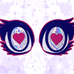 May include: Embroidered patches featuring stylized eyes with a heart-shaped pupil. The patches have a dark blue background with a pink outline and white and silver details. The eye's pupil is a bright pink heart shape.