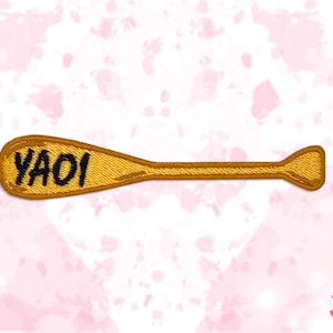 May include: A gold-colored embroidered patch in the shape of an oar. The word "YAOI" is embroidered in black letters on the paddle. The patch has a gold border and is set against a pink and white background.