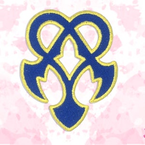 May include: Embroidered patch with a blue and gold design. The patch has a symmetrical shape with a central diamond and curved elements. The design is outlined in gold thread, creating a contrasting border.