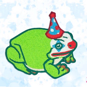 May include: Embroidered patch of a green frog with a clown face and a party hat. The frog has a red nose, blue and white eye makeup, and a red and blue party hat. The patch has a stitched texture.