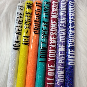 May include: Set of eight glitter pens with different colored glitter and black text. The text on the pens reads: "ACA-BELIEVE IT", "ACA-SCUSE ME", "CRUSHED IT", "I DON'T SPEAK IDSER", "I LOVE YOU AWESOME NERDS", "DON'T PUT ME DOWN FOR CARDIO", "DIXIE CHICKS SERIOUS".