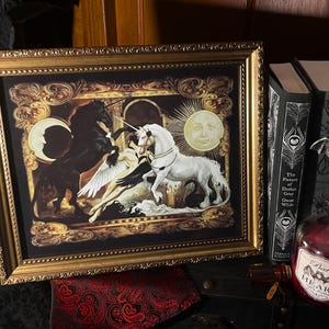 May include: A framed artwork with a gold ornate frame, depicting a black unicorn and a white unicorn with a winged figure. A full moon and a sun with a face are in the background. Beside the frame are two books, a bottle with a bat stopper.