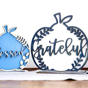 Puede incluir: Dos letreros de madera con forma de calabaza con las palabras "Blessed" y "Grateful" recortadas en el centro. El letrero "Blessed" está pintado de azul y el letrero "Grateful" es blanco.