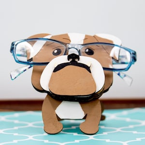 Puede incluir: Figura de bulldog de madera con gafas de montura azul. El bulldog es marrón y blanco con una nariz negra y un hocico blanco. Las gafas tienen lentes transparentes. La figura está sobre una superficie estampada en azul y blanco.