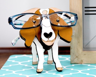 May include: A wooden dog-shaped eyeglass holder with blue-rimmed glasses. The dog is white, brown, and tan, with a black heart-shaped nose. A clock with Roman numerals is in the background. The holder is on a turquoise patterned surface.