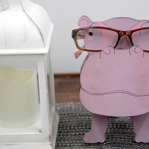 Lavender painted wood standing Hippo figurine. Larger Mouth / nose sticks out to hold glasses. Brown glasses on purple hippos face. Light pink cute wavy mouth, nostrils, & ears, black eyes white pupils. Standing on a desk next to lantern to show size