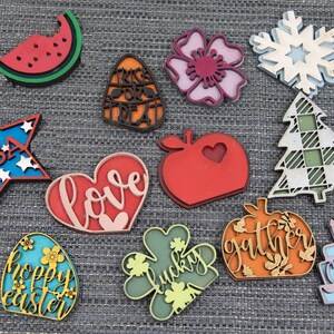 Puede incluir: Una colecci&oacute;n de 12 adornos de madera cortados con l&aacute;ser que presentan varios temas y dise&ntilde;os. Los adornos incluyen una sand&iacute;a, un ma&iacute;z dulce, una flor, un copo de nieve, un &aacute;rbol, un coraz&oacute;n con la palabra "love", una manzana, una calabaza con la palabra "gather", un pastel con la palabra "celebrate", una estrella con la palabra "USA", un tr&eacute;bol de cuatro hojas con la palabra "lucky" y un huevo con las palabras "happy easter".