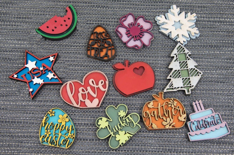 Puede incluir: Una colecci&oacute;n de 12 adornos de madera cortados con l&aacute;ser que presentan varios temas y dise&ntilde;os. Los adornos incluyen una sand&iacute;a, un ma&iacute;z dulce, una flor, un copo de nieve, un &aacute;rbol, un coraz&oacute;n con la palabra "love", una manzana, una calabaza con la palabra "gather", un pastel con la palabra "celebrate", una estrella con la palabra "USA", un tr&eacute;bol de cuatro hojas con la palabra "lucky" y un huevo con las palabras "happy easter".
