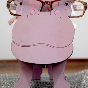 Lavender painted wood standing Hippo figurine. Larger Mouth / nose sticks out to hold glasses. Brown glasses on purple hippos face. Light pink cute wavy mouth, nostrils, and ears, black eyes white pupils. Standing on a desk & sparkly silver placemat
