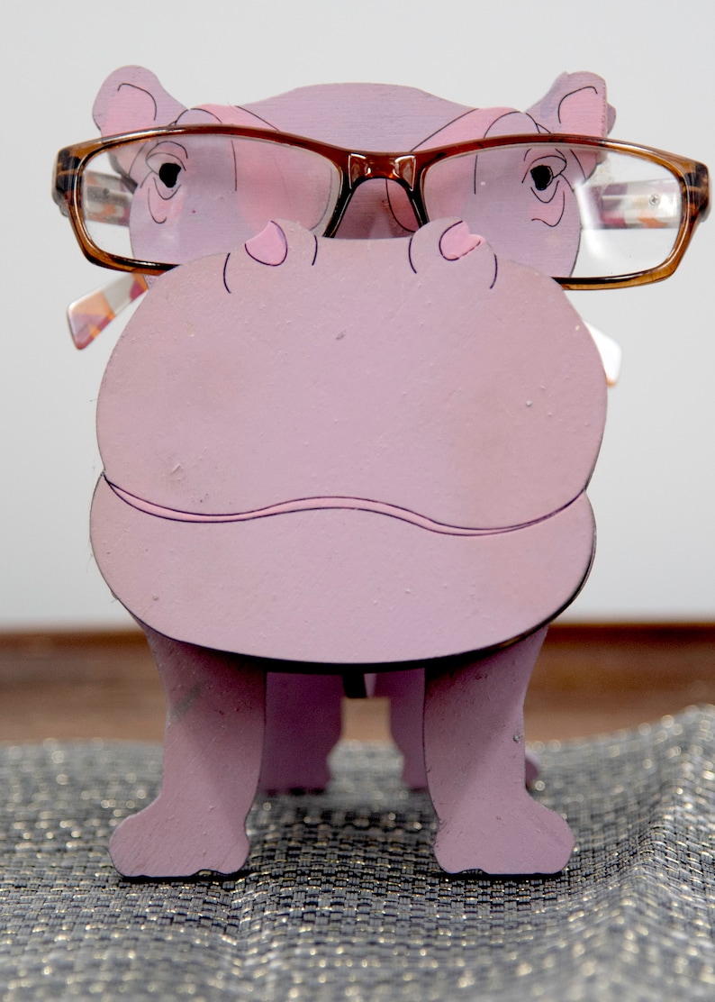 Lavender painted wood standing Hippo figurine. Larger Mouth / nose sticks out to hold glasses. Brown glasses on purple hippos face. Light pink cute wavy mouth, nostrils, and ears, black eyes white pupils. Standing on a desk & sparkly silver placemat