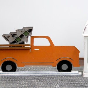 Puede incluir: Cami&oacute;n de madera naranja con un &aacute;rbol de Navidad a cuadros en la caja. El cami&oacute;n est&aacute; sobre un soporte plateado y est&aacute; al lado de una linterna blanca con una vela dentro.