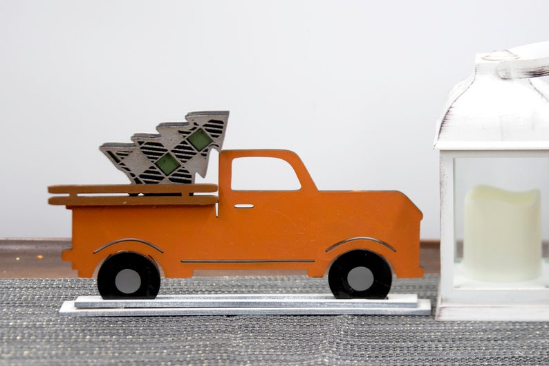 Puede incluir: Cami&oacute;n de madera naranja con un &aacute;rbol de Navidad a cuadros en la caja. El cami&oacute;n est&aacute; sobre un soporte plateado y est&aacute; al lado de una linterna blanca con una vela dentro.