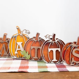 May include: Five wooden pumpkins with the word "WATTS" spelled out in the center of each pumpkin. The pumpkins are painted in shades of orange and brown with a glittery finish. The pumpkins are sitting on a plaid tablecloth.