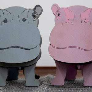 Grey & lavender wooden standing Hippos. Mouth / nose sticks out to hold glasses. Darker Grey cute wavy mouth, nostrils, & ears. Black eyes white pupils lavender Hippo has pink mouth, nostrils, eyes & ears. Standing on a desk & silver sparkly placemat
