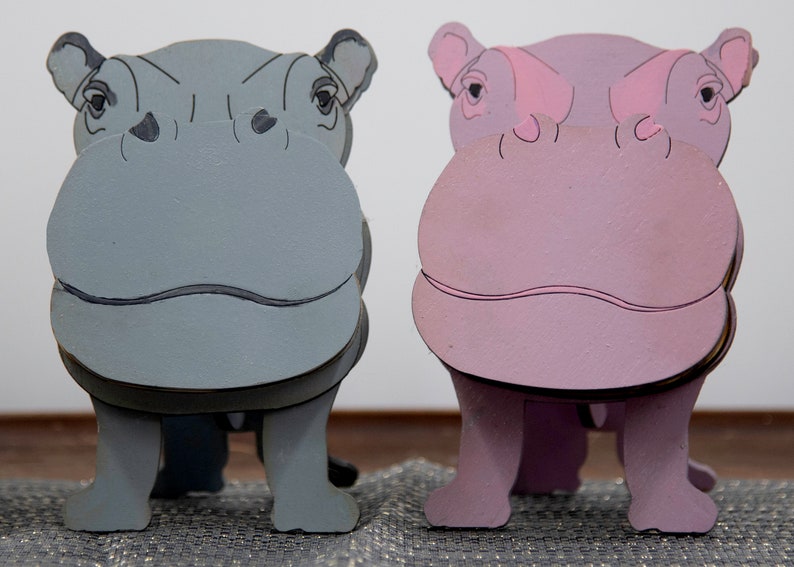 Grey & lavender wooden standing Hippos. Mouth / nose sticks out to hold glasses. Darker Grey cute wavy mouth, nostrils, & ears. Black eyes white pupils lavender Hippo has pink mouth, nostrils, eyes & ears. Standing on a desk & silver sparkly placemat