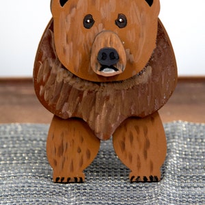 Wooden brown grizzley bear. Brown body & head, darker steaks for fur, Darker brown chest & light streaks for texture. protruding nose to hold glasses, black smaller nose & mouth, black eyes white pupils. sitting on a desk by large clock for size.