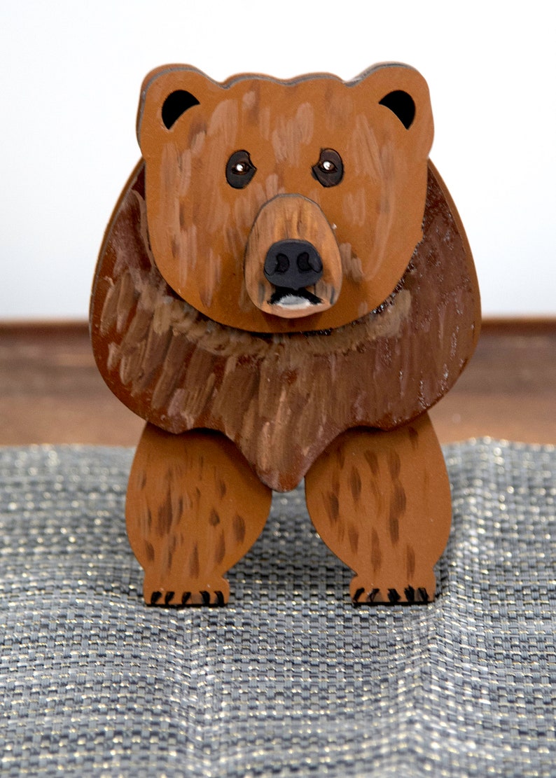 Wooden brown grizzley bear. Brown body & head, darker steaks for fur, Darker brown chest & light streaks for texture. protruding nose to hold glasses, black smaller nose & mouth, black eyes white pupils. sitting on a desk by large clock for size.