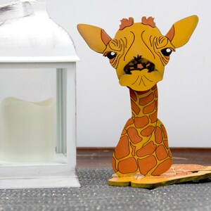 May include: A wooden giraffe figurine with a yellow and orange patterned body. The giraffe has a long neck and a sad expression. The figurine is sitting on a gray surface next to a white lantern.