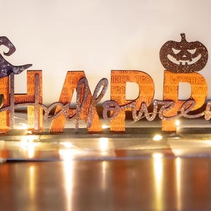 Puede incluir: Decoración de Halloween de madera con las palabras "HAPPY Halloween" en naranja y purpurina. Un gato negro y un sombrero de bruja están a la izquierda, y una calabaza a la derecha. La decoración está iluminada con pequeñas luces.