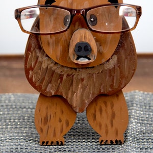 Wooden brown grizzley bear- brown glasses on its face. Brown body & head, darker steaks for fur, Darker brown chest & light streaks for texture. protruding nose holds glasses, black nose & mouth, black eyes white pupils. On desk on silver placemat