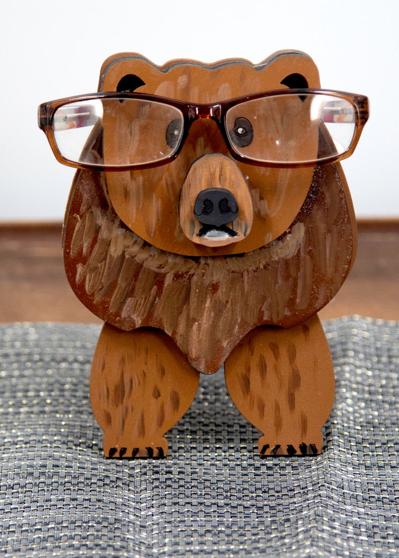 Wooden brown grizzley bear- brown glasses on its face. Brown body & head, darker steaks for fur, Darker brown chest & light streaks for texture. protruding nose holds glasses, black nose & mouth, black eyes white pupils. On desk on silver placemat