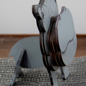 Side view of light grey painted wood standing Hippo figurine. Larger Mouth/ nose sticks out to hold glasses. Rounded body, little tail darker Grey cute wavy mouth,nostrils, & ears, black eyes white pupils. Standing on a desk & sparkly silver placemat