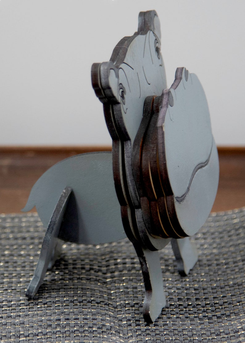 Side view of light grey painted wood standing Hippo figurine. Larger Mouth/ nose sticks out to hold glasses. Rounded body, little tail darker Grey cute wavy mouth,nostrils, & ears, black eyes white pupils. Standing on a desk & sparkly silver placemat