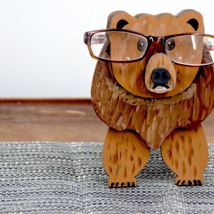 Wooden brown grizzley bear- brown glasses on its face. Brown body & head, darker steaks for fur, Darker brown chest & light streaks for texture. protruding nose holds glasses, black nose & mouth, black eyes white pupils. On desk on silver placemat