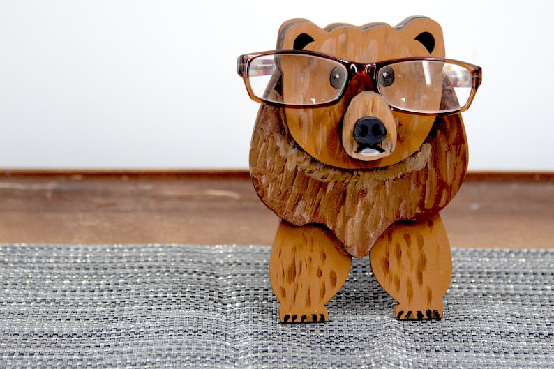Wooden brown grizzley bear- brown glasses on its face. Brown body & head, darker steaks for fur, Darker brown chest & light streaks for texture. protruding nose holds glasses, black nose & mouth, black eyes white pupils. On desk on silver placemat