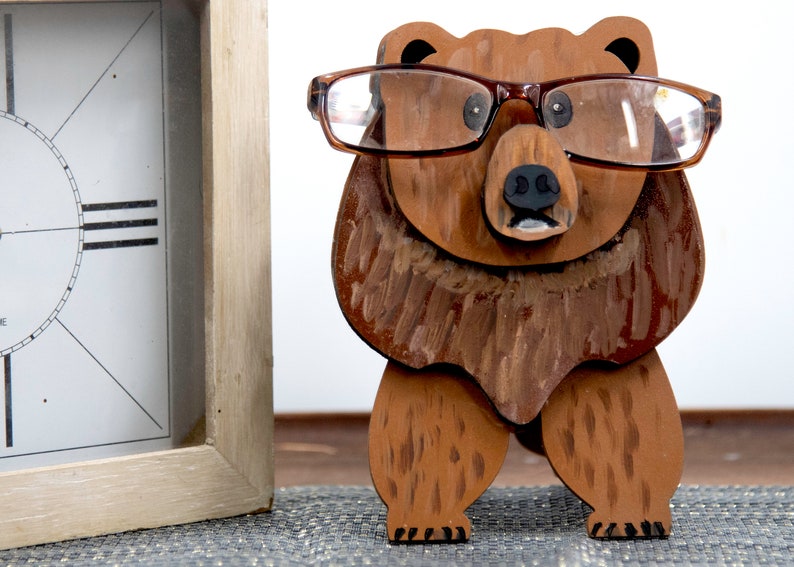 Wooden brown grizzley bear- glasses on its face. Brown body & head, darker steaks for fur, Darker brown chest & light streaks for texture. protruding nose holds glasses, black nose & mouth, black eyes white pupils. On desk by large clock for size.