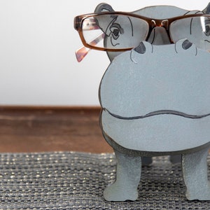 Light grey painted wood standing Hippo figurine. Larger Mouth / nose sticks out to hold glasses. Brown glasses on Grey hippos face. darker Grey cute wavy mouth, nostrils, and ears, black eyes white pupils. Standing on a desk & sparkly silver placemat