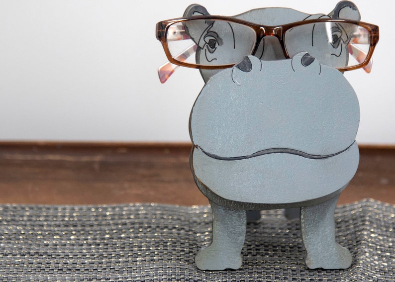 Light grey painted wood standing Hippo figurine. Larger Mouth / nose sticks out to hold glasses. Brown glasses on Grey hippos face. darker Grey cute wavy mouth, nostrils, and ears, black eyes white pupils. Standing on a desk & sparkly silver placemat
