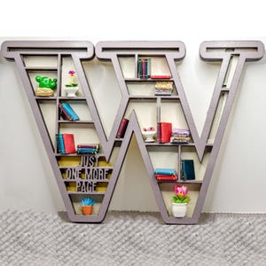 May include: A wooden bookshelf shaped like the letter W with shelves filled with miniature books and small potted plants. The text "Just One More Page" is written on the front of the bookshelf.
