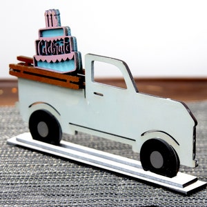 Puede incluir: Una camioneta de madera blanca con una cama de madera marr&oacute;n que lleva un pastel de cumplea&ntilde;os rosa y azul con la palabra "Celebrate" en ella. La camioneta est&aacute; sobre una base de madera blanca.