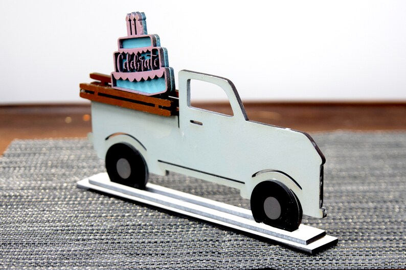Puede incluir: Una camioneta de madera blanca con una cama de madera marr&oacute;n que lleva un pastel de cumplea&ntilde;os rosa y azul con la palabra "Celebrate" en ella. La camioneta est&aacute; sobre una base de madera blanca.
