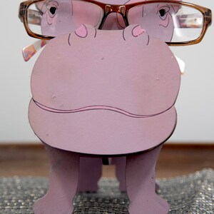 Lavender painted wood standing Hippo figurine. Larger Mouth / nose sticks out to hold glasses. Brown glasses on purple hippos face. Light pink cute wavy mouth, nostrils, and ears, black eyes white pupils. Standing on a desk & sparkly silver placemat