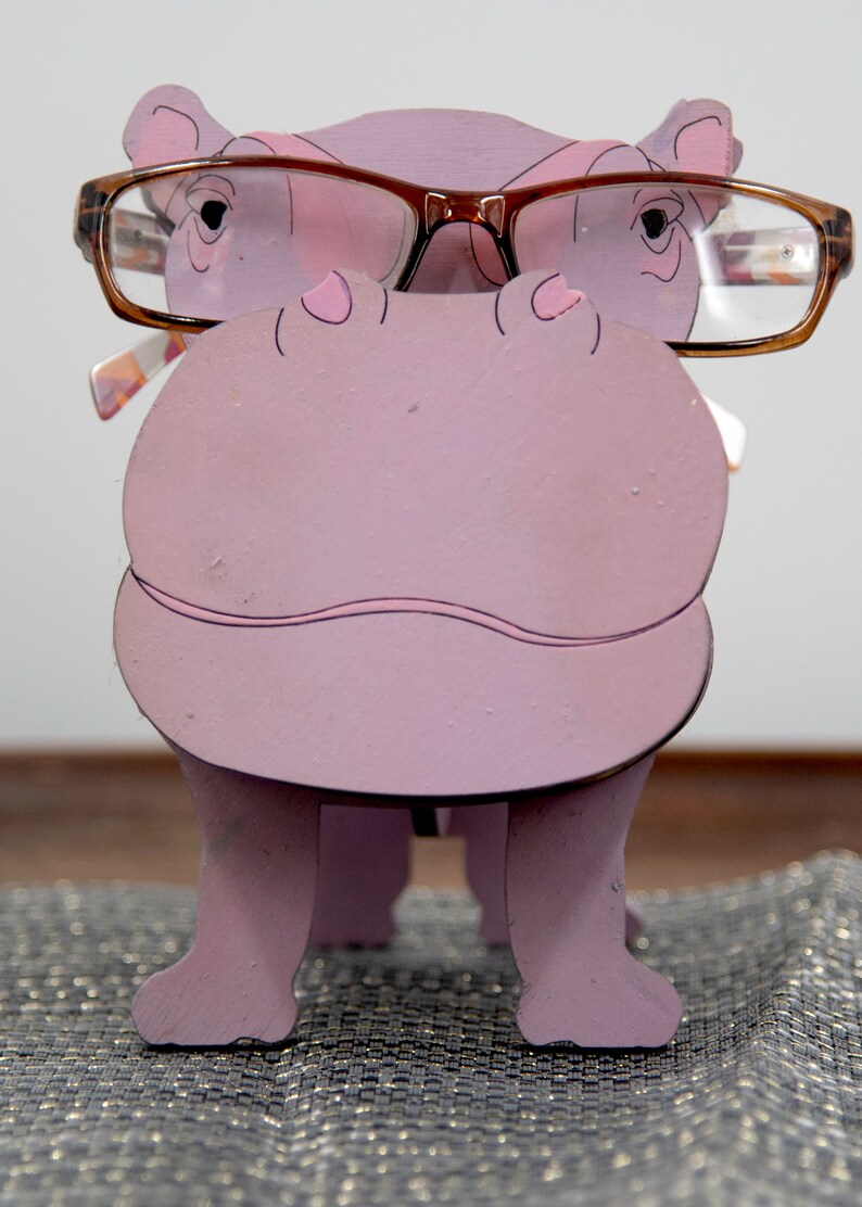Lavender painted wood standing Hippo figurine. Larger Mouth / nose sticks out to hold glasses. Brown glasses on purple hippos face. Light pink cute wavy mouth, nostrils, and ears, black eyes white pupils. Standing on a desk & sparkly silver placemat