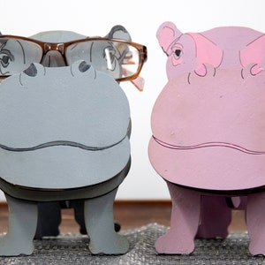 Grey and lavender wooden standing Hippos. Mouth / nose sticks out to hold glasses. Brown glasses on Grey hippos face. darker Grey mouth, nostrils, eyes and ears. lavender Hippo has pink mouth, nostrils, eyes & ears. Both standing on a desk & placemat