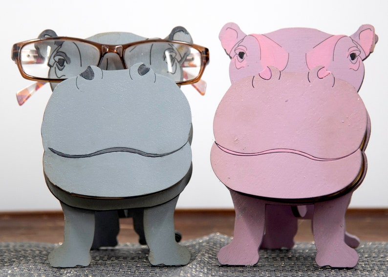 Grey and lavender wooden standing Hippos. Mouth / nose sticks out to hold glasses. Brown glasses on Grey hippos face. darker Grey mouth, nostrils, eyes and ears. lavender Hippo has pink mouth, nostrils, eyes & ears. Both standing on a desk & placemat