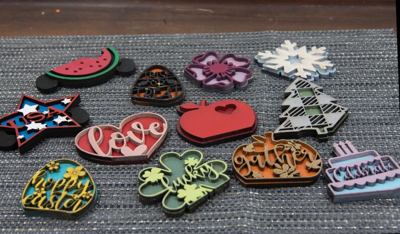 Puede incluir: Una colecci&oacute;n de 12 adornos de madera cortados con l&aacute;ser con varios dise&ntilde;os y colores. Los adornos presentan temas como el amor, la suerte, la reuni&oacute;n, la celebraci&oacute;n, feliz pascua, EE. UU., truco o trato, sand&iacute;a, manzana, copo de nieve y una flor.