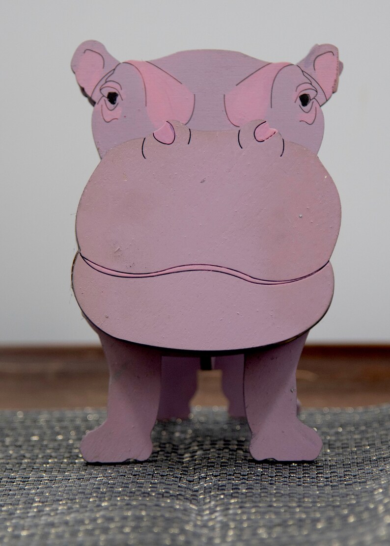 Lavender painted wood standing Hippo figurine. Larger Mouth / nose sticks out to hold glasses. Light pink cute wavy mouth, nostrils, & ears, black eyes white pupils. Cute rounded body and stubby tail. Standing on a desk & silver sparkly placemat