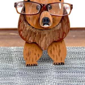 Wooden brown grizzley bear- brown glasses on its face. Brown body & head, darker steaks for fur, Darker brown chest & light streaks for texture. protruding nose holds glasses, black nose & mouth, black eyes white pupils. On desk on silver placemat