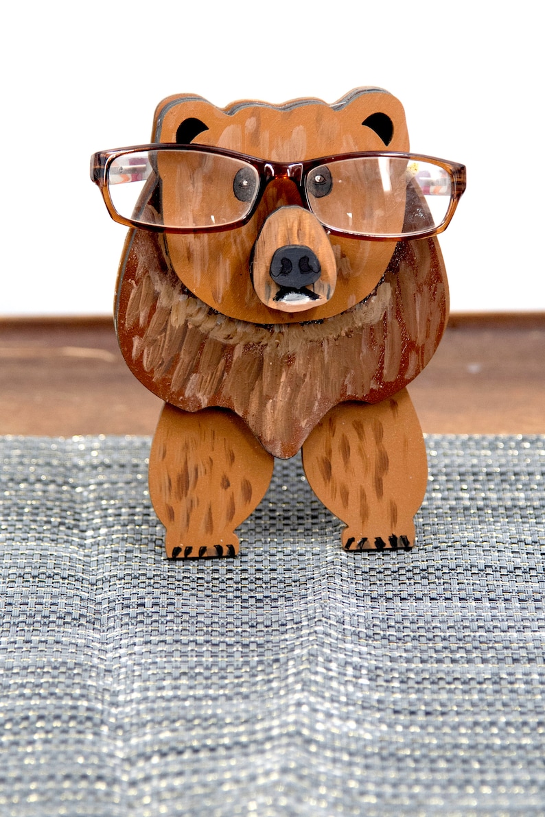 Wooden brown grizzley bear- brown glasses on its face. Brown body & head, darker steaks for fur, Darker brown chest & light streaks for texture. protruding nose holds glasses, black nose & mouth, black eyes white pupils. On desk on silver placemat