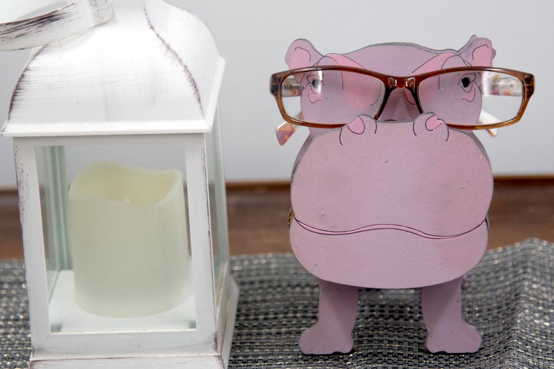Lavender painted wood standing Hippo figurine. Larger Mouth / nose sticks out to hold glasses. Brown glasses on purple hippos face. Light pink cute wavy mouth, nostrils, & ears, black eyes white pupils. Standing on a desk next to lantern to show size