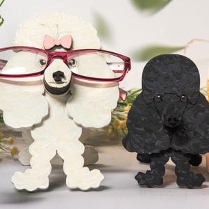 Puede incluir: Dos figuras de caniche de madera, una blanca y otra negra, están una al lado de la otra. El caniche blanco lleva gafas rosas y un lazo rosa. El caniche negro tiene un pelaje texturizado. Una vela y vegetación en el fondo.